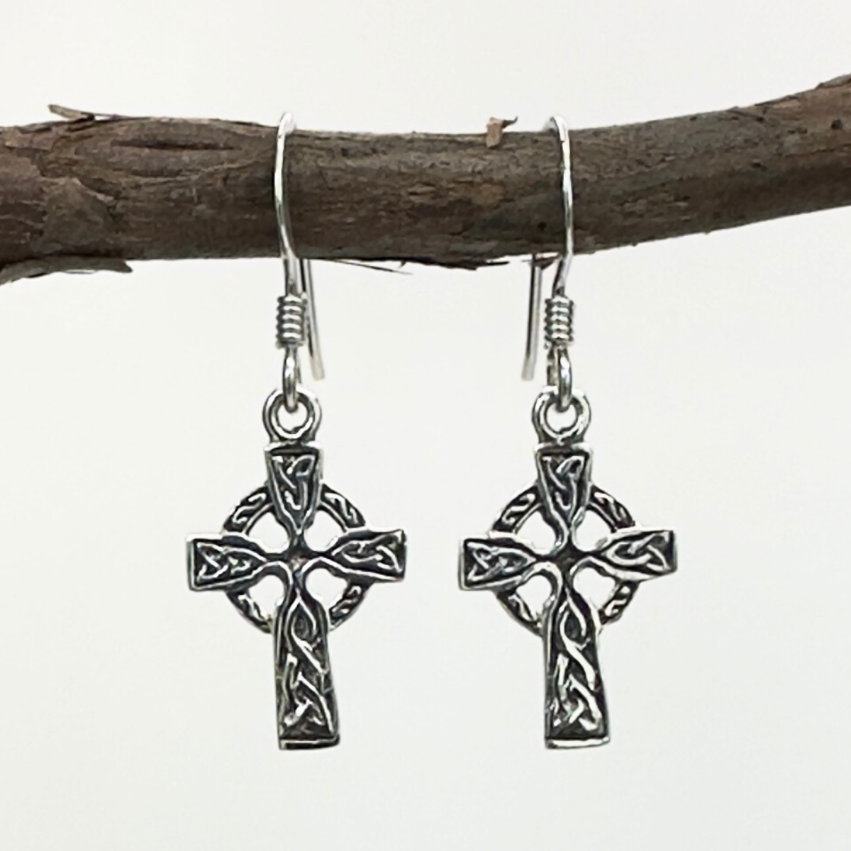 Silver Celtic cross earrings hanging on a branch with a white background