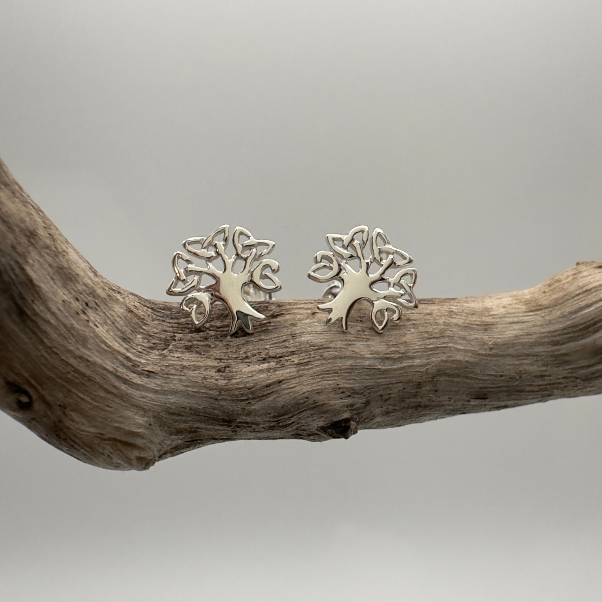 Pair of silver tree-shaped earrings on a wooden branch with a neutral background