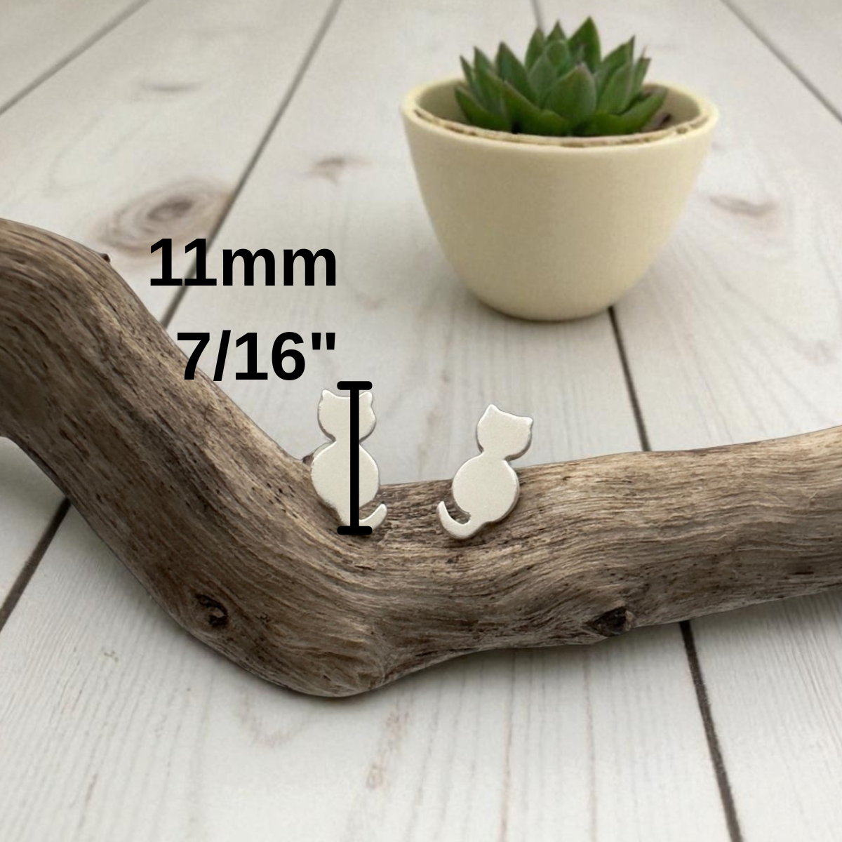 Small white cat-shaped earring on a wooden branch with measurement text and a potted plant in the background.