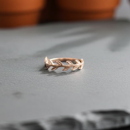 Rose gold leaf-patterned ring on a gray surface with blurred potted plants in the background