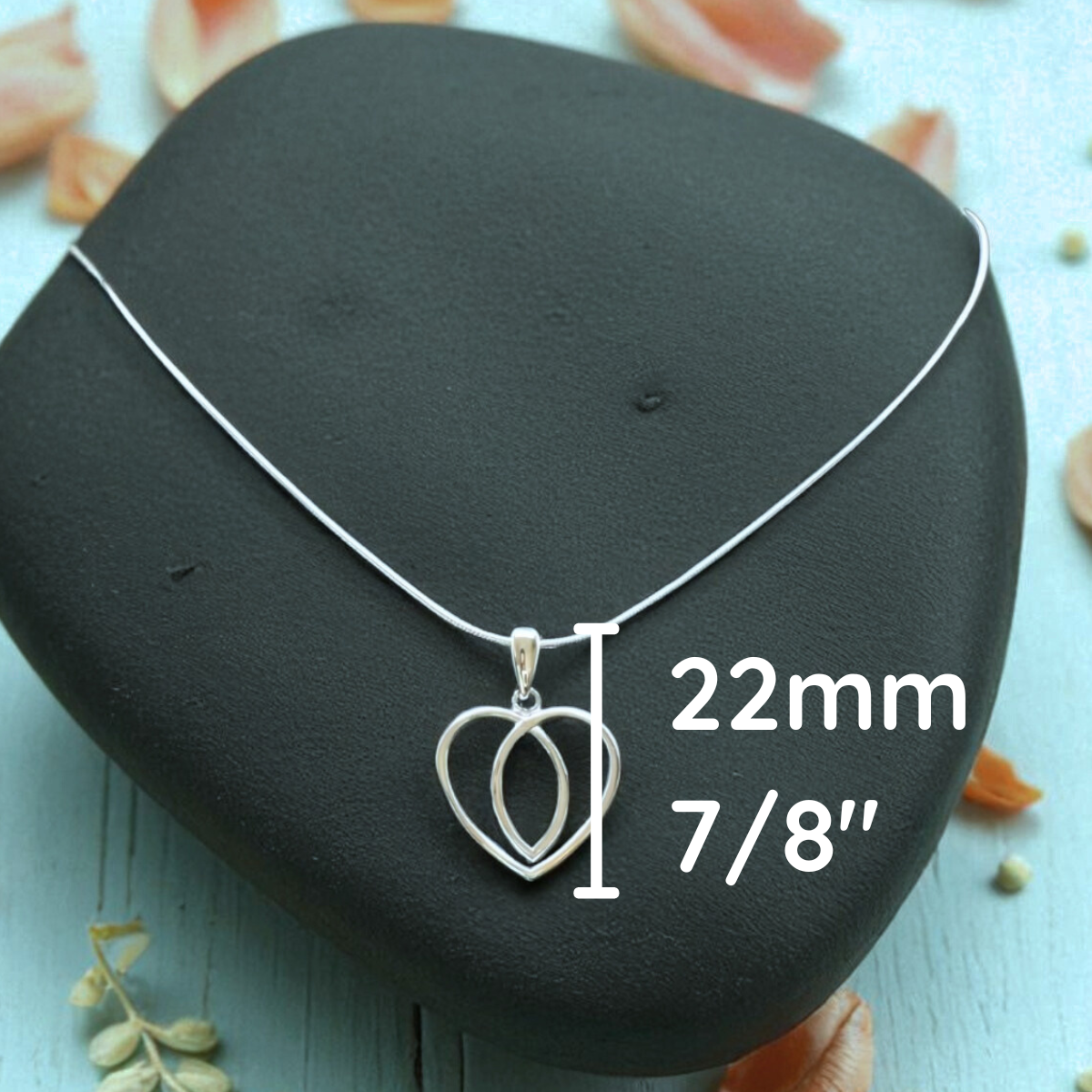 Silver necklace with heart pendant on a black stone, surrounded by dried flowers and leaves.